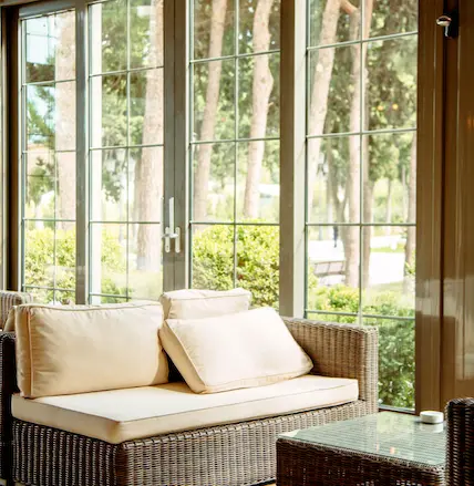 Floor-to-ceiling energy-efficient windows with colonial grids showcasing natural garden views, featuring a comfortable sunroom setting with wicker furniture and cream cushions